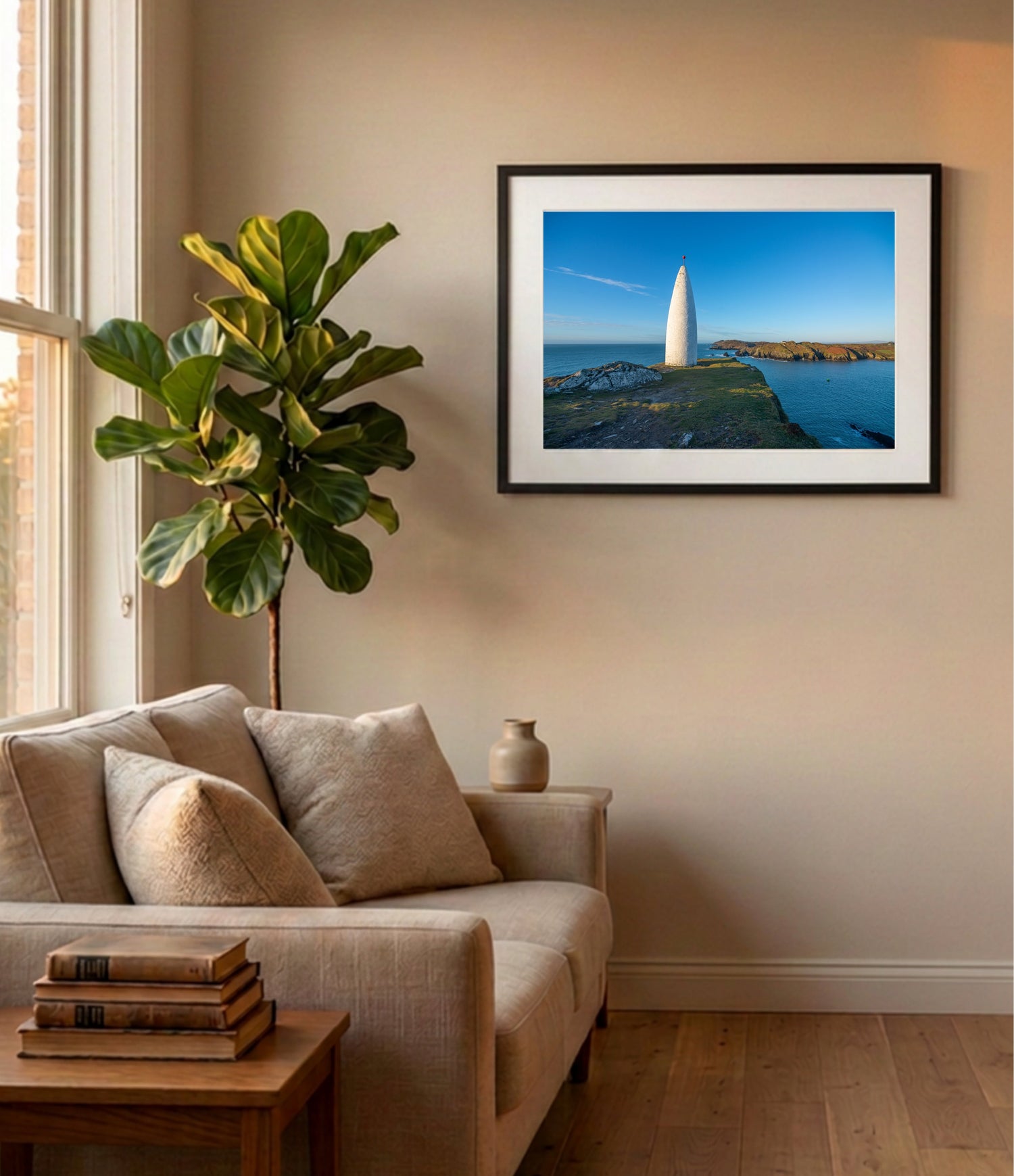A framed landscape photograph of a white lighthouse on a rocky coastline under a blue sky, displayed on a beige wall above a light beige sofa in a modern living room setting.